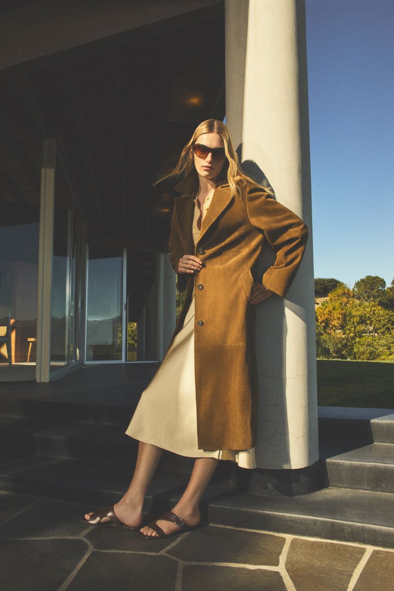 A fashionable adult female poses in a brown coat and sunglasses near a white column, set against an outdoor landscape with a clear blue sky.