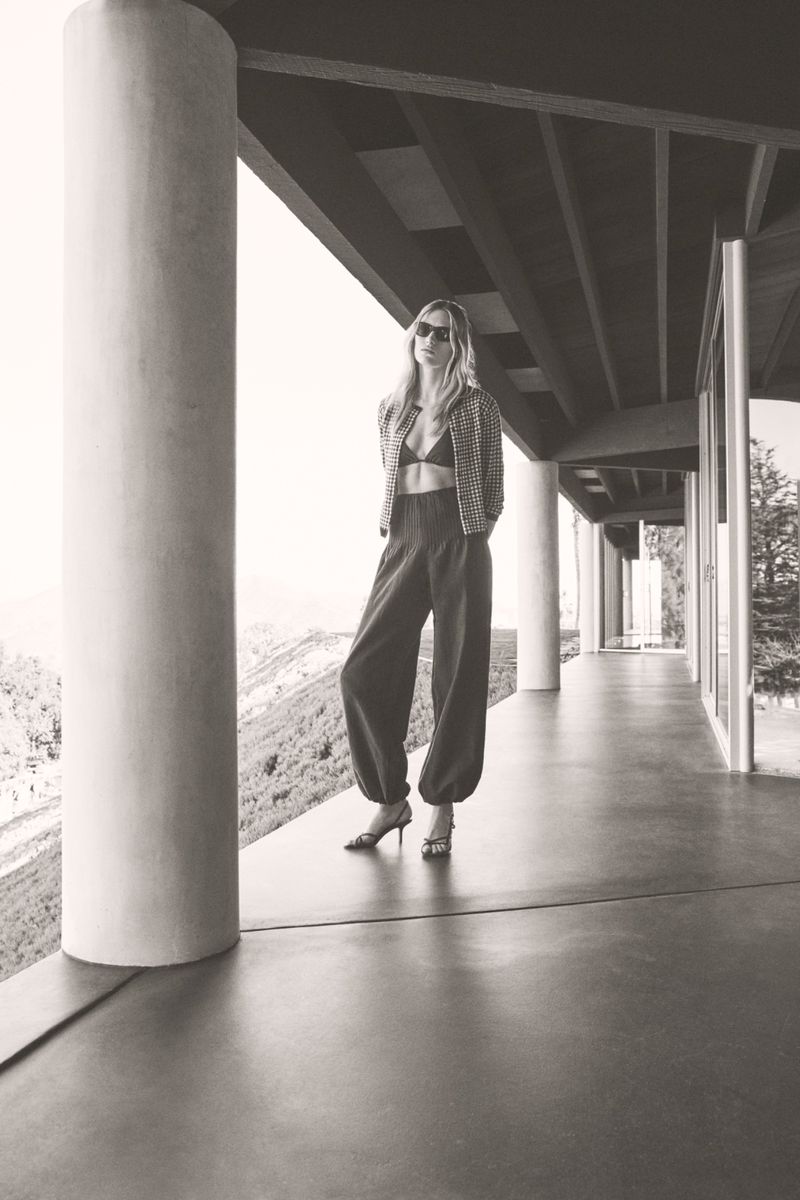 A monochrome full-body shot of a woman in stylish wide-leg trousers and a blazer, posing on a modern balcony with a mountainous view.