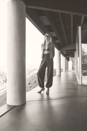A monochrome full-body shot of a woman in stylish wide-leg trousers and a blazer, posing on a modern balcony with a mountainous view.