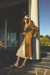 Model wearing a brown trench coat and cream skirt standing against a concrete pillar outdoors for Vince Spring 26.