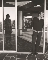 Monochrome fashion campaign image featuring a male model leaning in a doorway and a female model reflected in a glass pane.