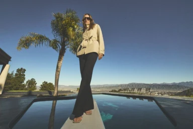Model standing on a diving board wearing a tan jacket and dark trousers by a pool with palm trees.