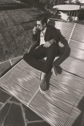 A man in a black suit relaxing on an outdoor lounger holding a small glass, photographed by Emma Summerton for Vince Spring 26.