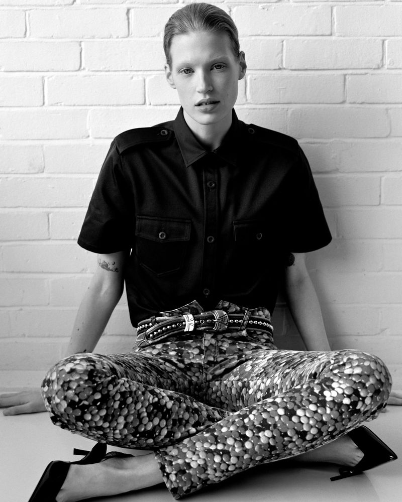 Monochrome portrait of a model sitting against a white brick wall wearing a short-sleeved buttoned shirt and patterned trousers