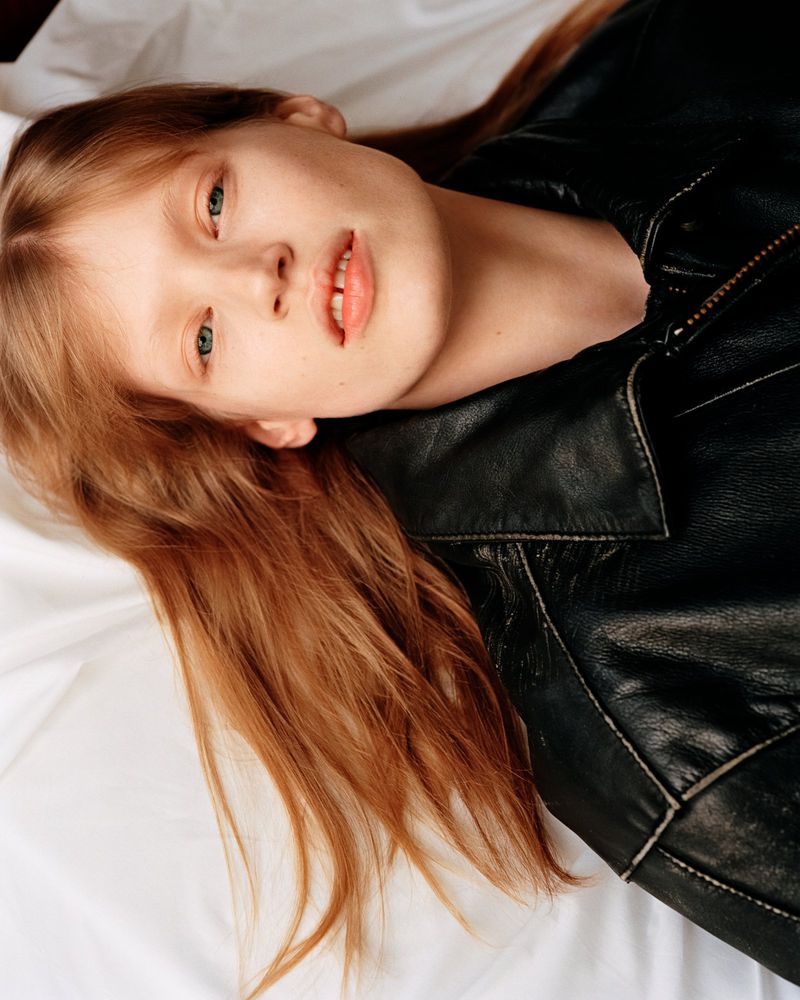 Close-up portrait of a young woman with red hair in a black leather jacket, lying on a white surface, creating a relaxed, stylish fashion image.