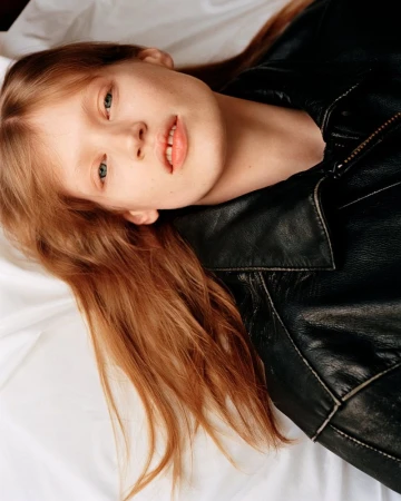 Close-up portrait of a young woman with red hair in a black leather jacket, lying on a white surface, creating a relaxed, stylish fashion image.