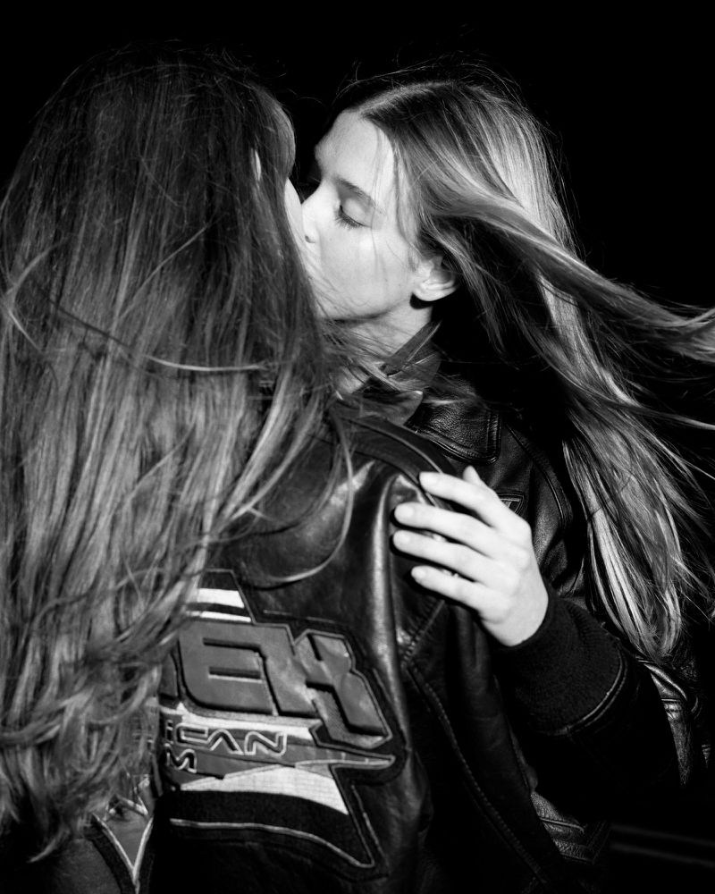 Monochrome portrait of two young women kissing, both with long hair, wearing leather jackets, against a dark, blurred background.