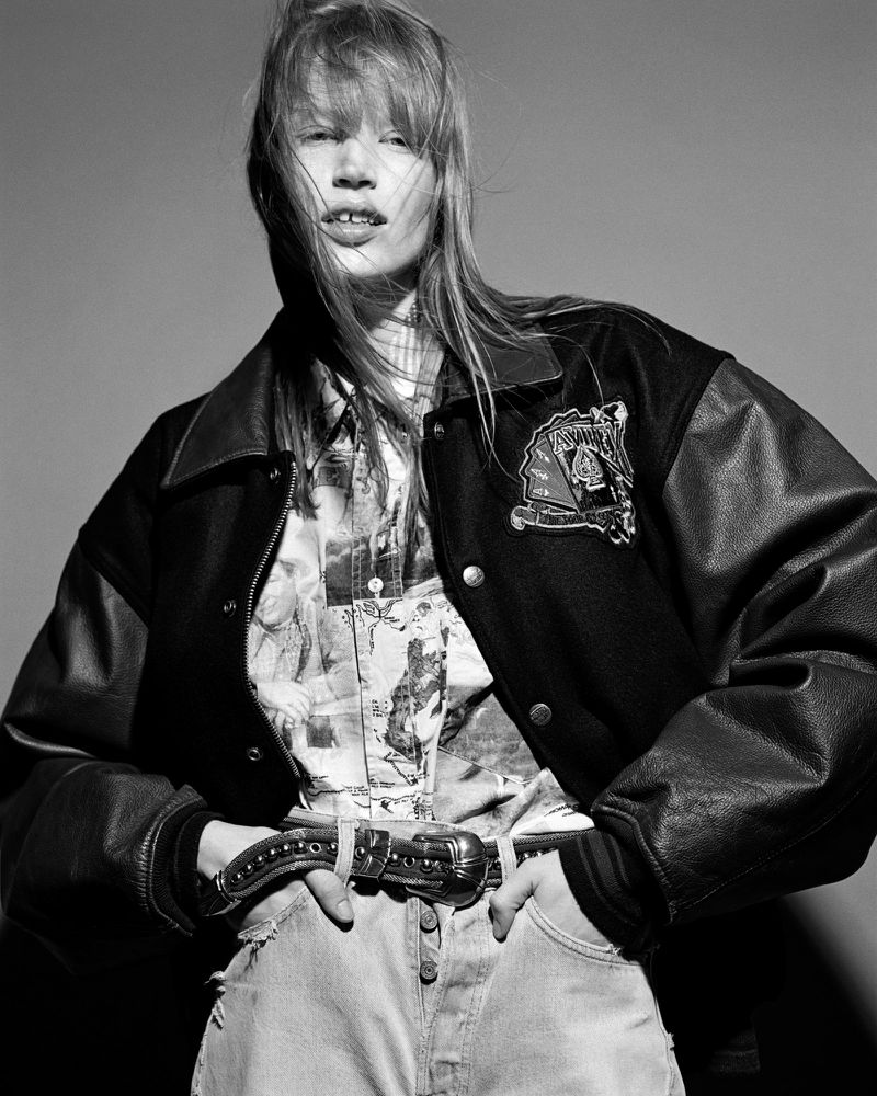 Black and white portrait of a young woman wearing a jacket, patterned blouse, jeans, and belt, hands in pockets, neutral background.