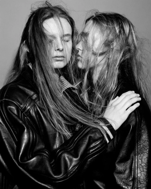 Two young women in black leather jackets, hair partially obscuring faces, pose intimately in a monochrome fashion-style photo studio portrait.