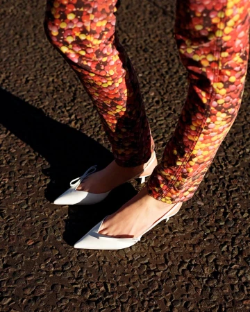 Close-up of a woman's legs wearing patterned pants and white kitten heel shoes on an asphalt surface, shot in natural sunlight.
