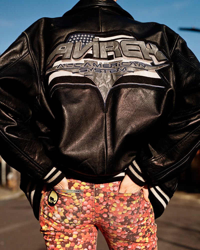 Person from the back in a black leather jacket with a logo on the back and patterned pants stands on an asphalt street.