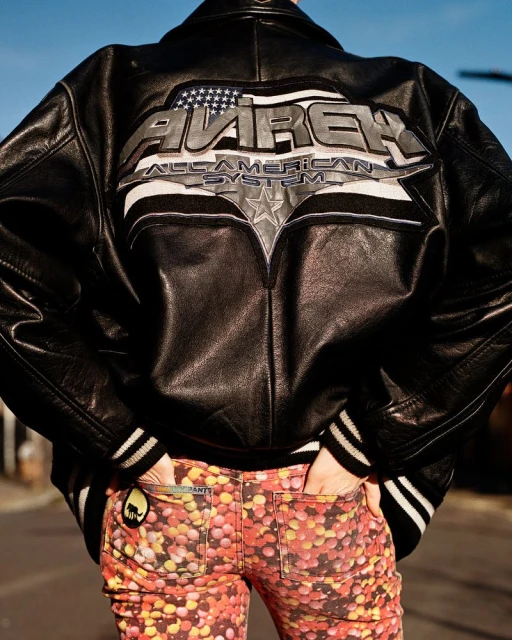 Person from the back in a black leather jacket with a logo on the back and patterned pants stands on an asphalt street.