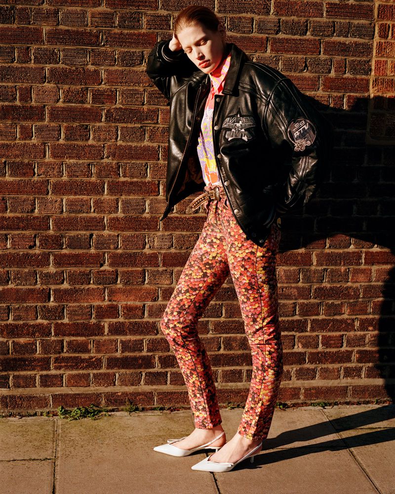 A fashionable young woman in patterned pants and black jacket leans against a brick wall outdoors on a sunny day, showcasing an editorial fashion style.