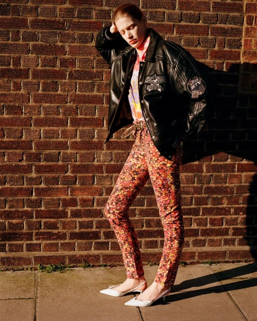 A fashionable young woman in patterned pants and black jacket leans against a brick wall outdoors on a sunny day, showcasing an editorial fashion style.