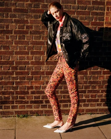 A fashionable young woman in patterned pants and black jacket leans against a brick wall outdoors on a sunny day, showcasing an editorial fashion style.
