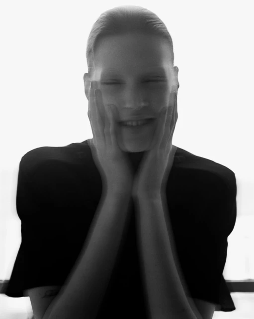 Monochrome portrait of a smiling woman with hands on her face, creating a blurred, dreamlike effect against a bright white background.