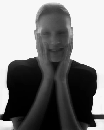 Monochrome portrait of a smiling woman with hands on her face, creating a blurred, dreamlike effect against a bright white background.