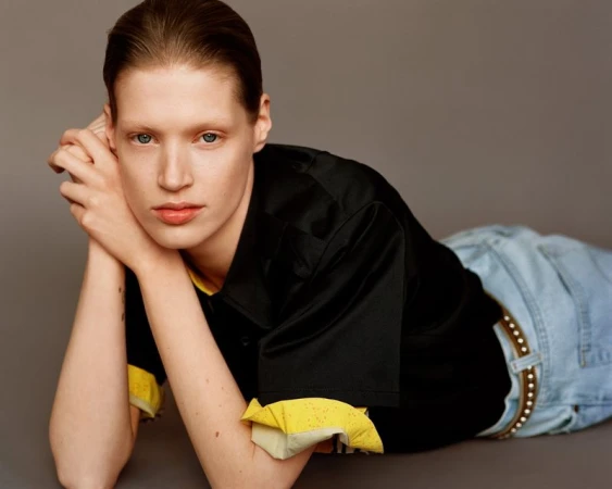 Model with slicked-back hair wearing a black shirt and blue jeans reclining against a neutral grey studio background