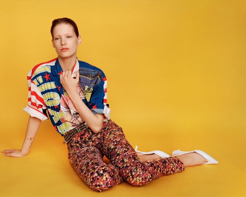 Woman in patterned pants and graphic top poses against a yellow backdrop in a fashion studio shot with natural lighting.