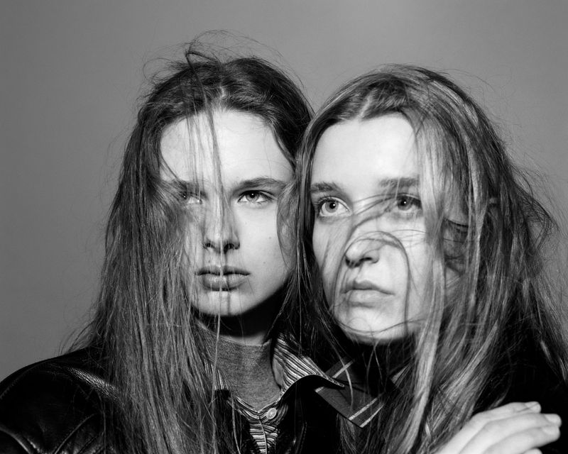 Black and white portrait of two fair-skinned young women with long hair, wearing leather jackets, posing in a photo studio against gray backdrop.