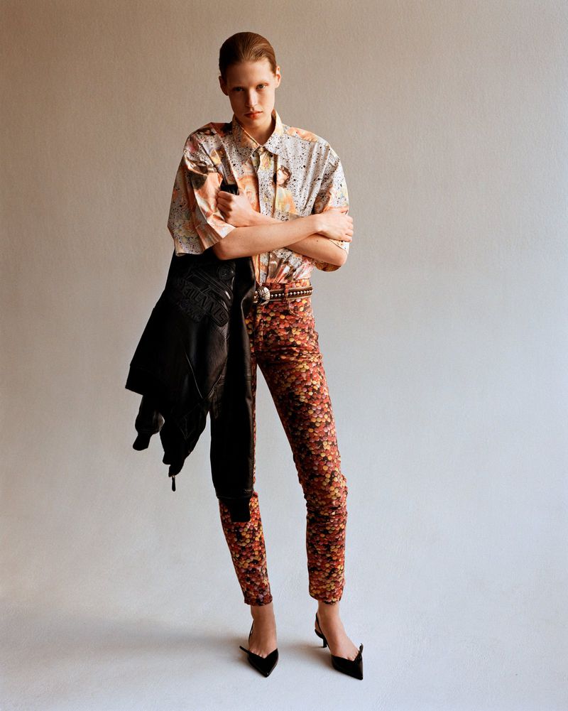 Fashion shot of a female model in printed shirt and pants, standing indoors with a leather jacket over her arm, posing confidently.