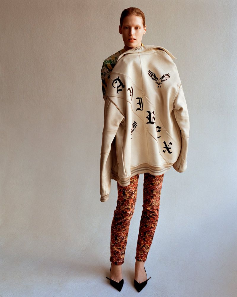 A slender fashion model poses in patterned leggings, a black bow heel, and an oversized cream bomber jacket against a plain backdrop.