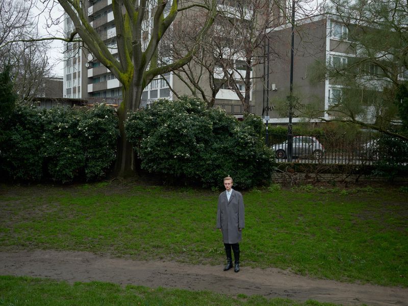 Full shot of an adult standing in an overcast outdoor scene with green grass, trees, and a tall apartment building in the background.