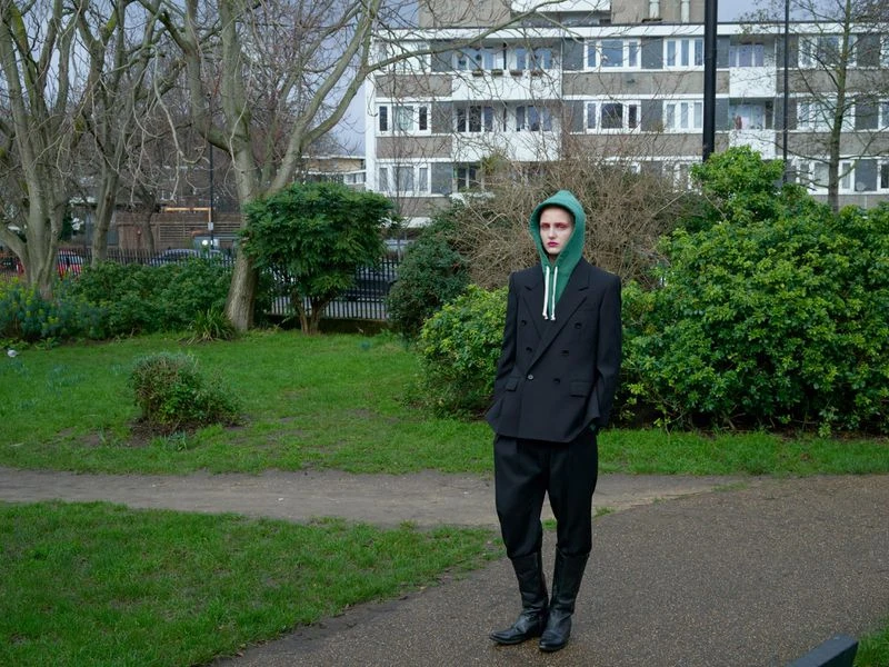 Person in black double-breasted jacket with green hooded sweatshirt stands in park setting, apartment building in background, overcast day.