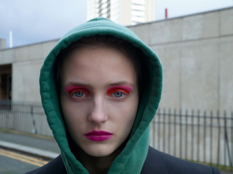 Close-up of a woman with bright makeup wearing a green hoodie against a concrete background, showcasing a modern and edgy beauty look.
