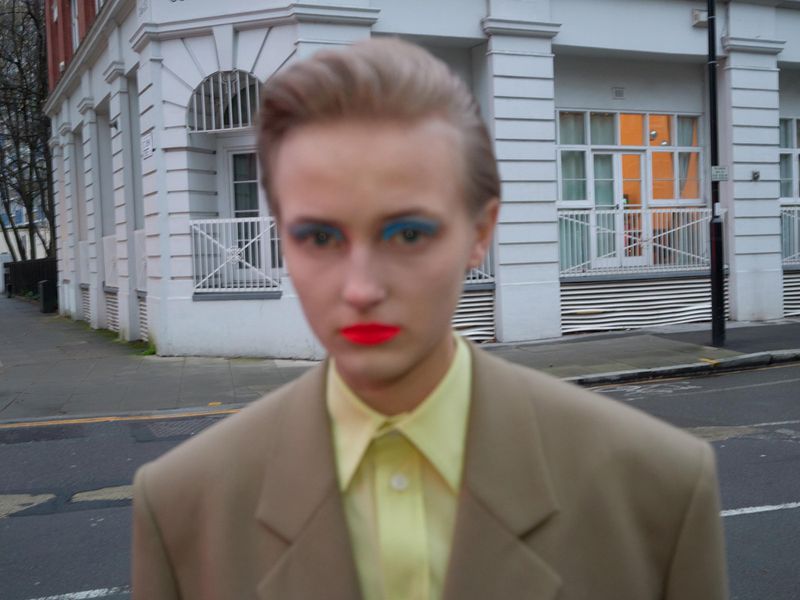 Close-up of person with brightly colored makeup posing in a beige suit on a city street with buildings in the background.