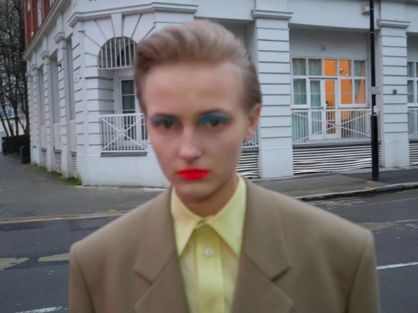 Close-up of person with brightly colored makeup posing in a beige suit on a city street with buildings in the background.