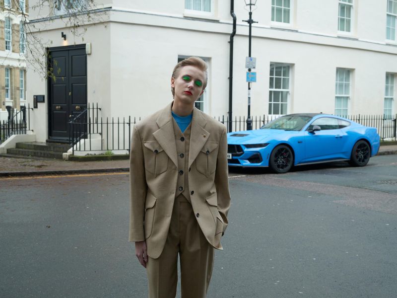 A person in a tan suit stands on a street near a bright blue sportscar and white buildings, creating a sophisticated and modern outdoor scene.