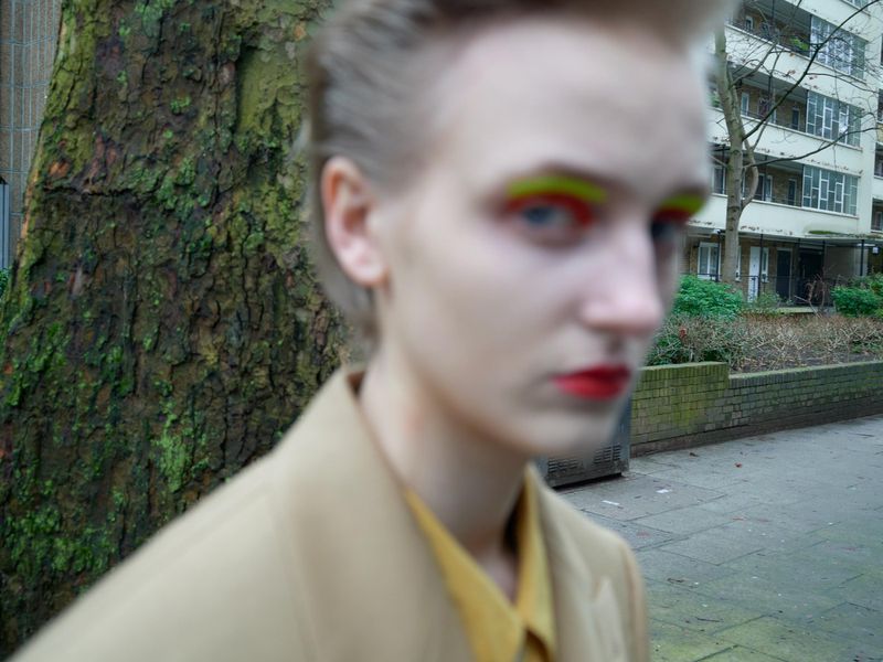 Close-up of a person with bright yellow and red makeup standing outside in front of a tree and residential buildings in an urban setting.