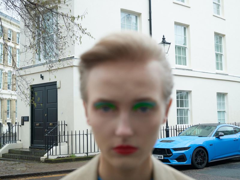 Close-up of a blurred model with colorful makeup and red lipstick, with a light-blue sports car in front of a white building.