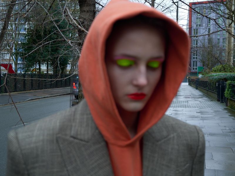 Woman in orange hoodie, blazer with bright eye makeup stands on urban street with brick buildings; fashion editorial shot.