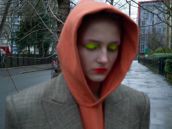 Woman in orange hoodie, blazer with bright eye makeup stands on urban street with brick buildings; fashion editorial shot. 