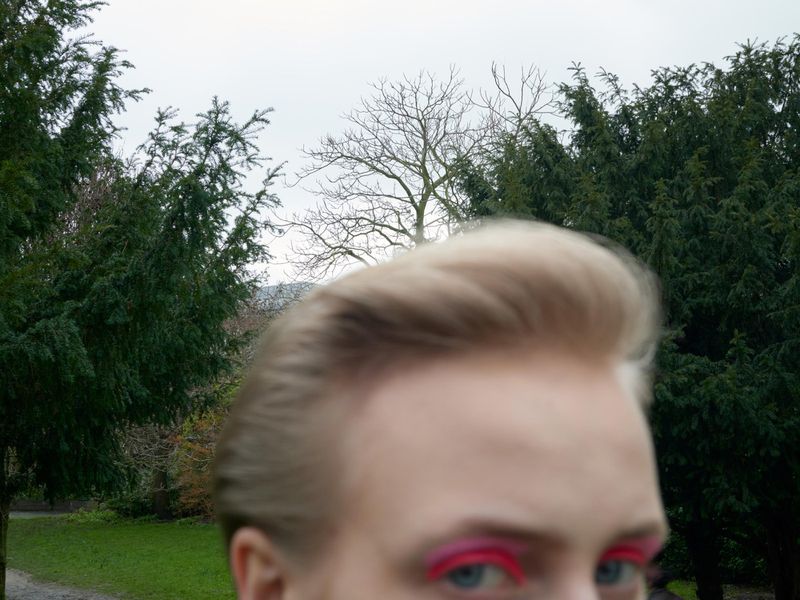 Close-up of a fair-skinned person with blond hair and bright pink eye makeup, looking directly at the camera in a park-like outdoor setting.