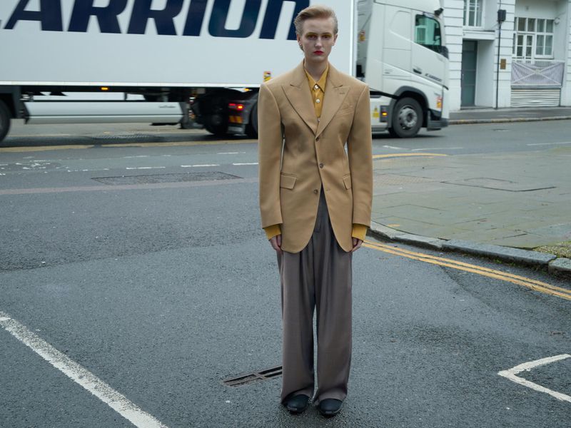 An adult stands on an urban street wearing a beige blazer, mustard shirt, and wide gray pants, posing near a large white truck on a city street.