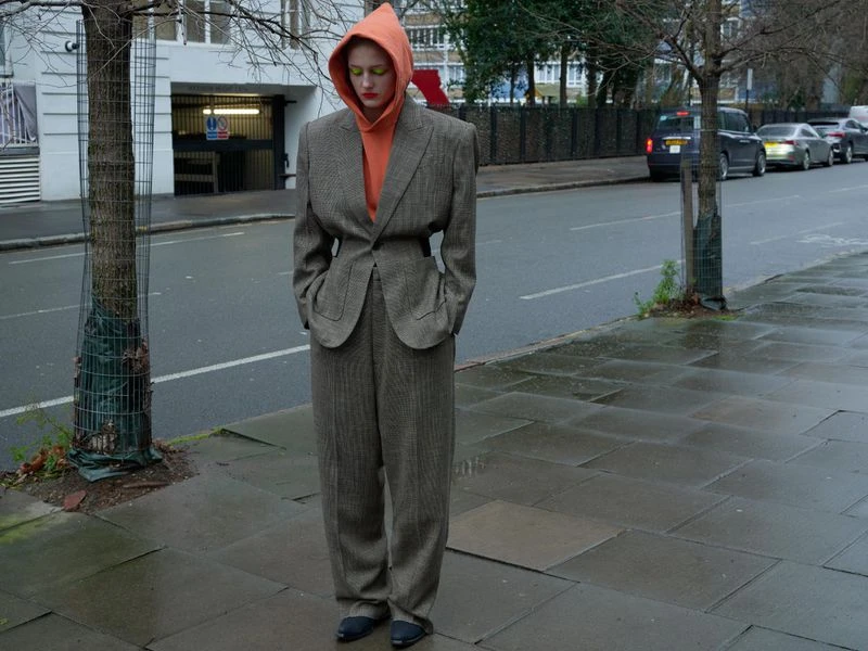 Adult in a brown herringbone suit and orange hooded sweatshirt poses on a city street with a building and cars in the background.