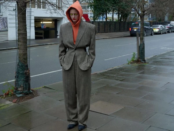 Adult in a brown herringbone suit and orange hooded sweatshirt poses on a city street with a building and cars in the background.