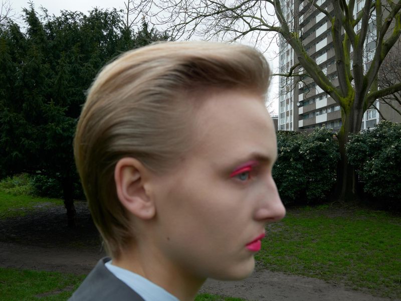 Close-up of a blonde-haired adult wearing pink makeup and a grey suit jacket, standing outdoors in a grassy urban park setting.
