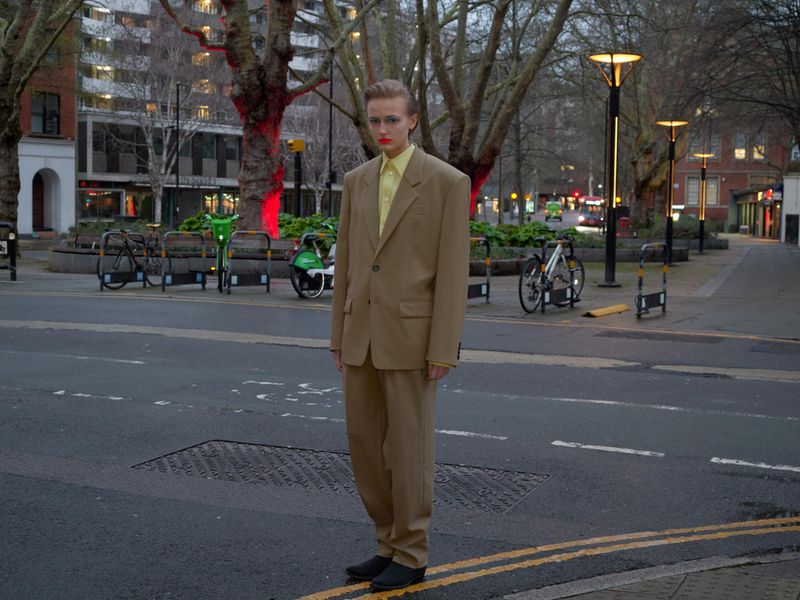 Fashionable adult stands on an urban street wearing a tan suit with a yellow shirt, black shoes and lipstick, with city buildings in the background.