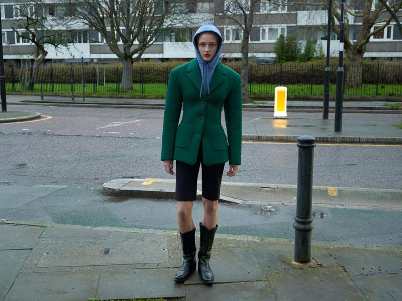 Woman in a green jacket and black shorts stands on a wet city sidewalk with apartment buildings and trees visible in the background.
