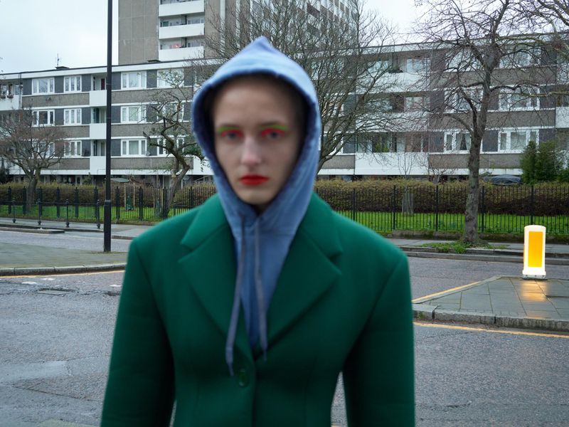 A person with green and red makeup stands on a city street wearing a blue hoodie and a green jacket, with buildings in the background.