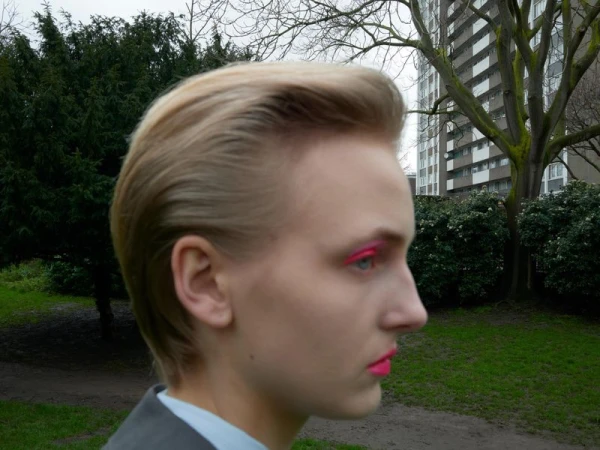 Profile portrait of a blonde male model with vibrant pink eye makeup outdoors in front of a residential tower block