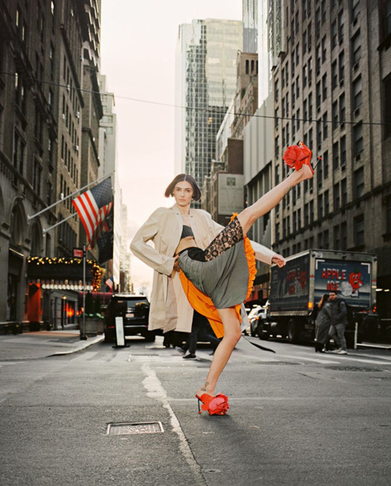 Fashion model in a beige trench coat and red heels performs a high kick on a city street with tall buildings and an American flag.