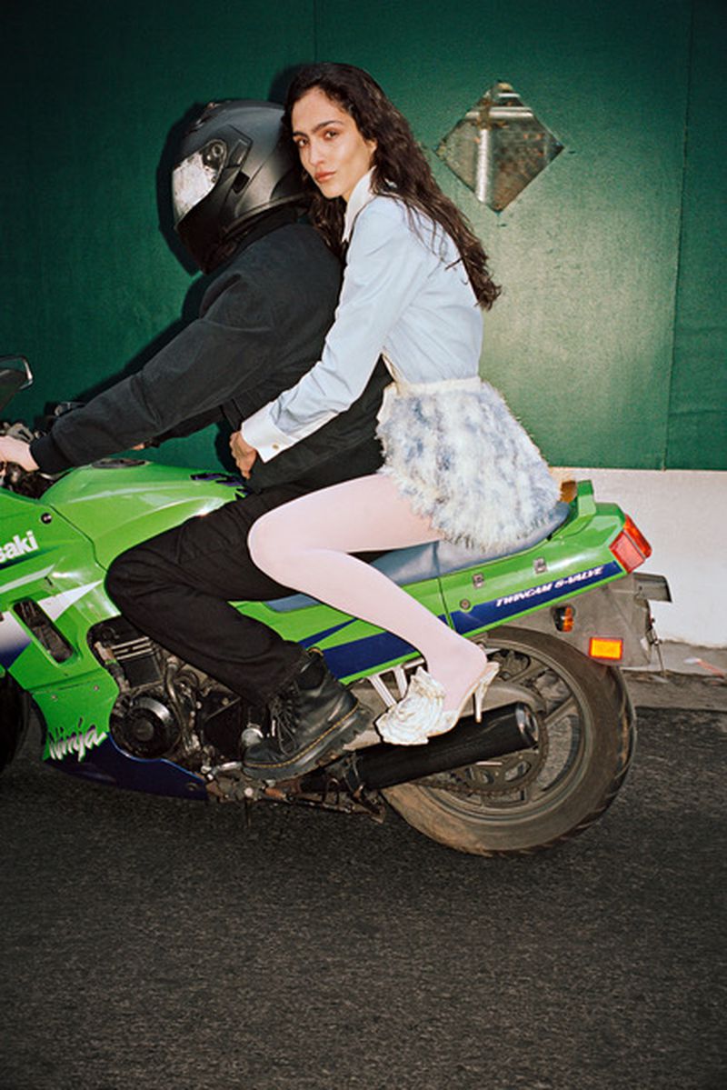 Model riding on the back of a green motorcycle driven by a person in a helmet against a green wall