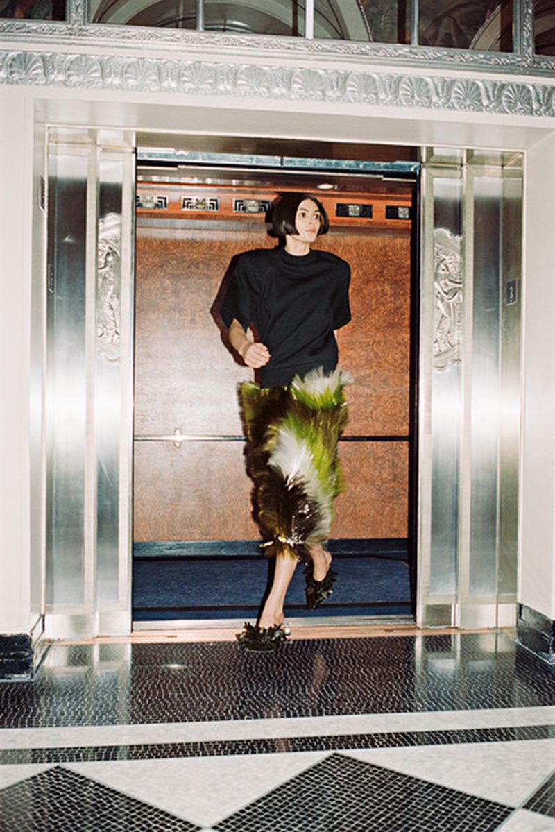 Fashionable woman exits an elevator in a black top and green feathered skirt on a patterned floor in an elegant building interior.