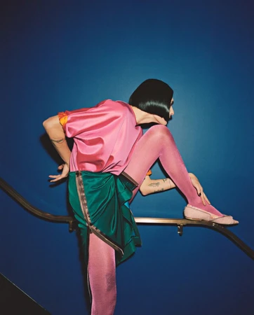 Woman in pink top and green skirt poses on metal railing against blue background, wearing pink tights and shoes, black bob hairstyle.