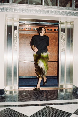Model exiting an elevator wearing a dark top and feathered skirt in an architectural hotel lobby.